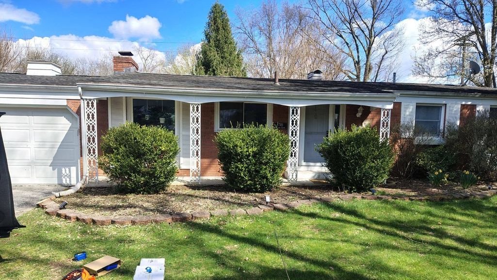 Three rounded shrubs trimmed in front of a brick house with a white garage door, and a leaf blower and hedge trimmer resting on a freshly mown lawn.