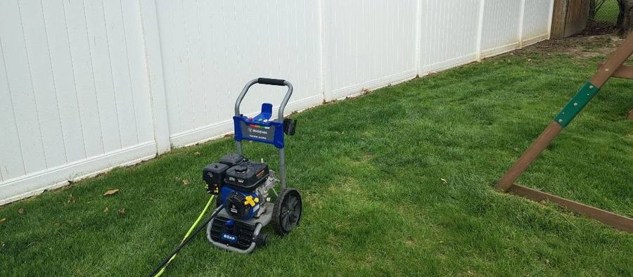 A gas pressure washer with a green hose standing on the grass beside a white vinyl privacy fence.